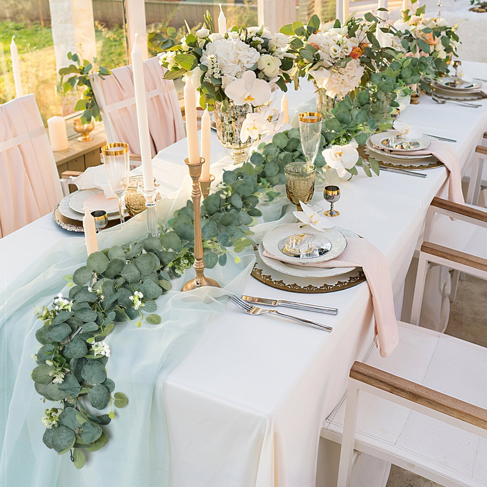 Artificial eucalyptus garland with white flowers draped across a formal dining table setting with place settings and candles.