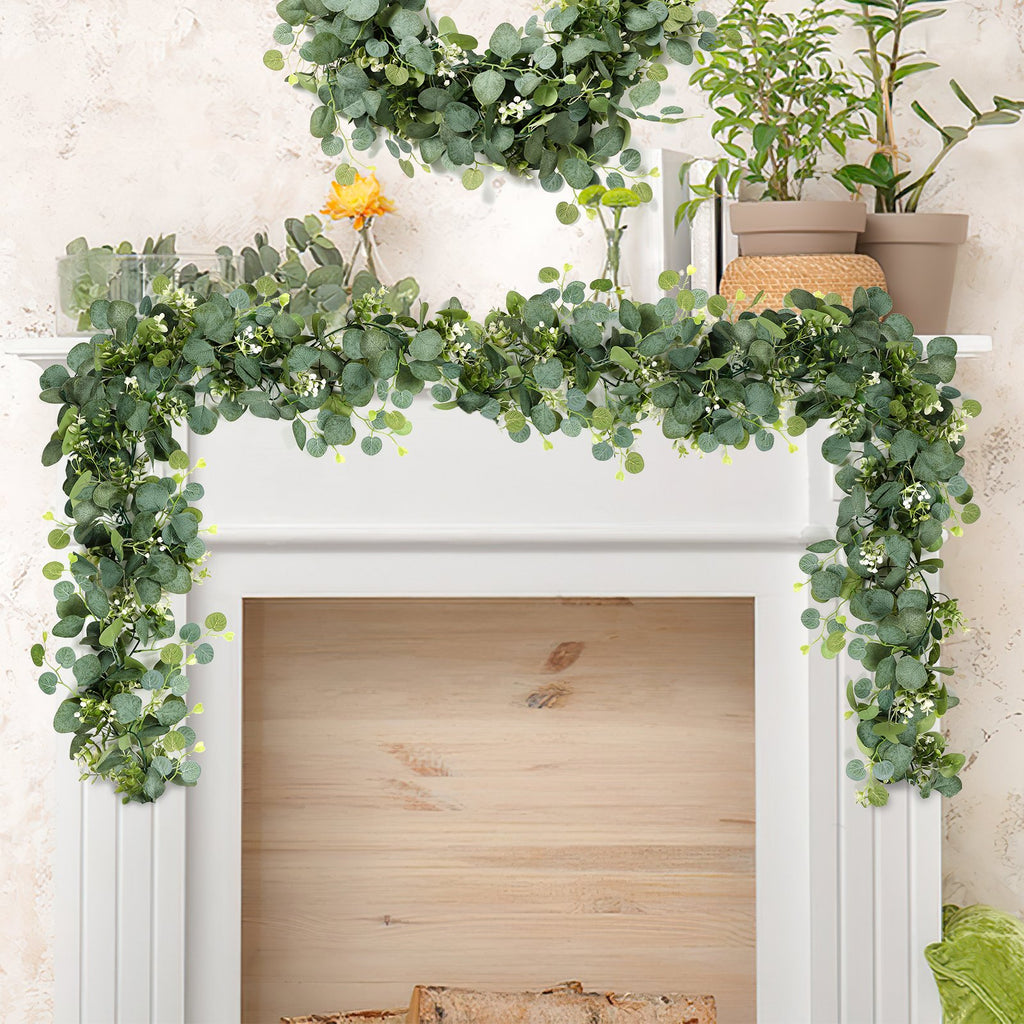 Artificial eucalyptus garland with white flowers draped over a white mantelpiece with plants and a vase with a yellow flower.