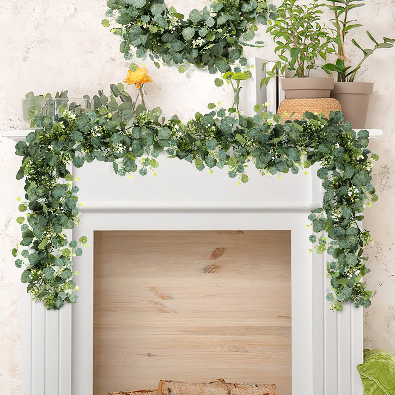 Artificial eucalyptus garland with white flowers draped over a white mantelpiece with plants and a vase with a yellow flower.