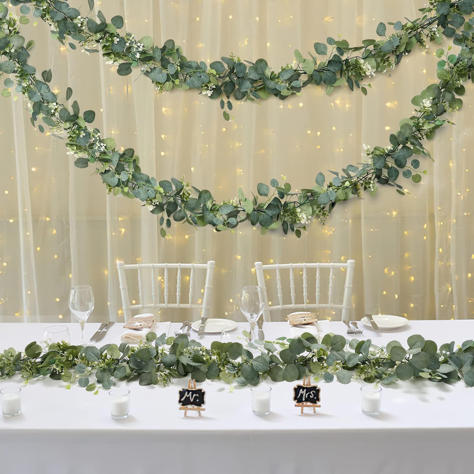 Artificial eucalyptus garland with white flowers draped on a table and against a backdrop with fairy lights.