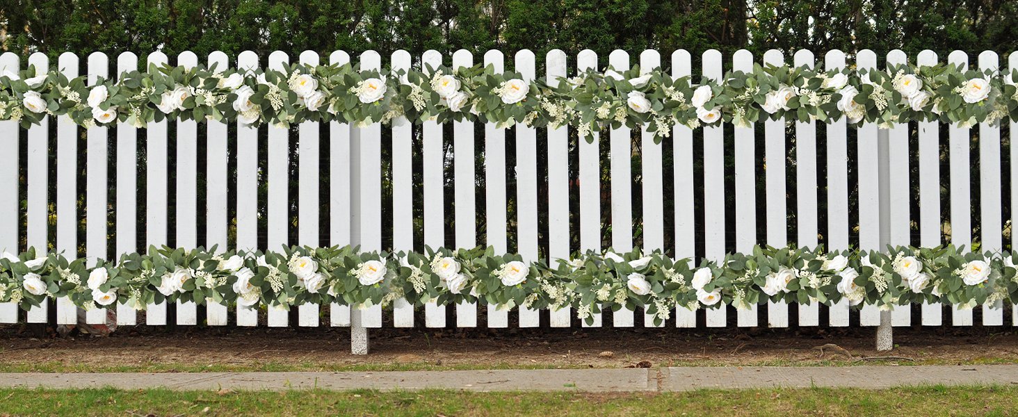 Two artificial eucalyptus garlands with white roses and seeded leaves draped on a white picket fence.