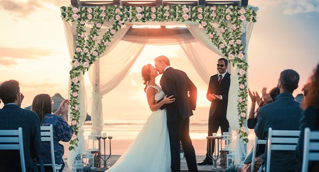 Wedding ceremony on a beach with a couple kissing under an arch decorated with artificial eucalyptus garland and pink roses.