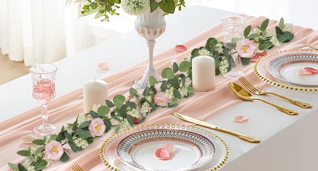 Artificial eucalyptus garland with pink roses and white flowers draped on a pink table runner with place settings.
