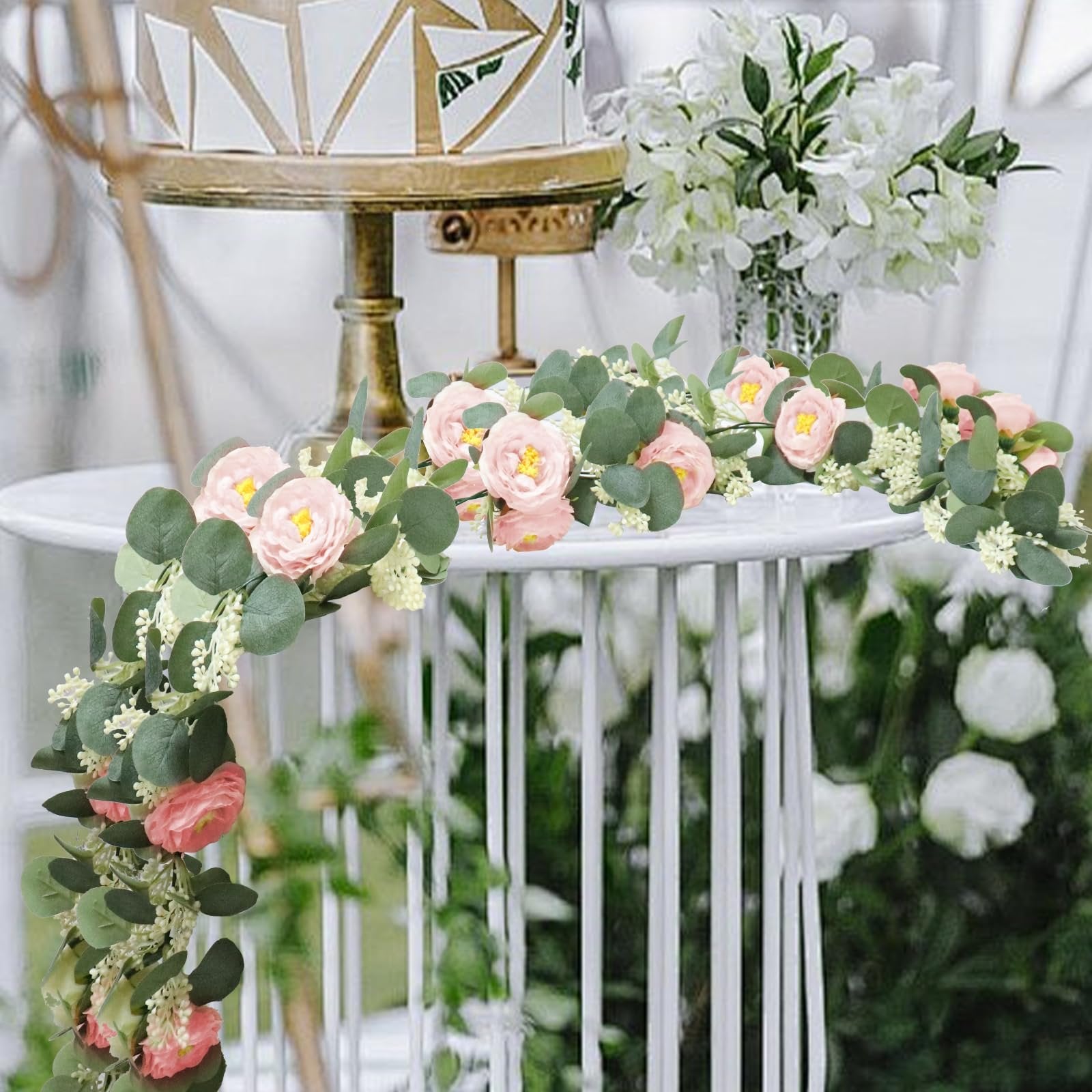 Artificial eucalyptus garland with pink roses and white gypsophila draped over a table with a cake stand and floral arrangement.