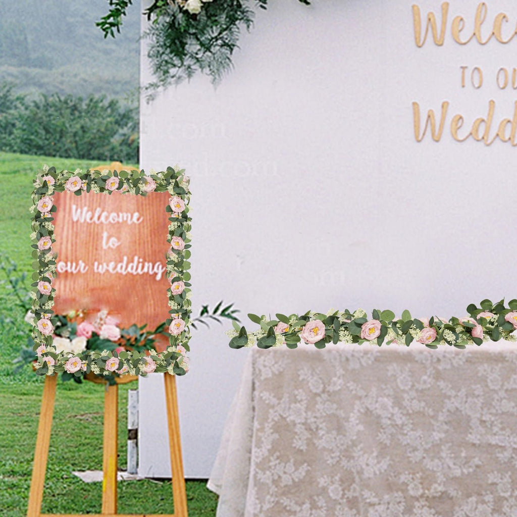 Artificial eucalyptus garland with pink roses and white flowers framing a welcome sign and draped on a table