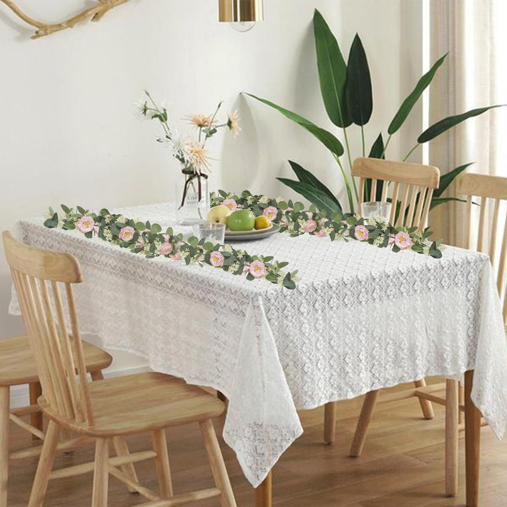 Artificial eucalyptus garland with pink roses and white gypsophila draped across a lace tablecloth on a dining table.