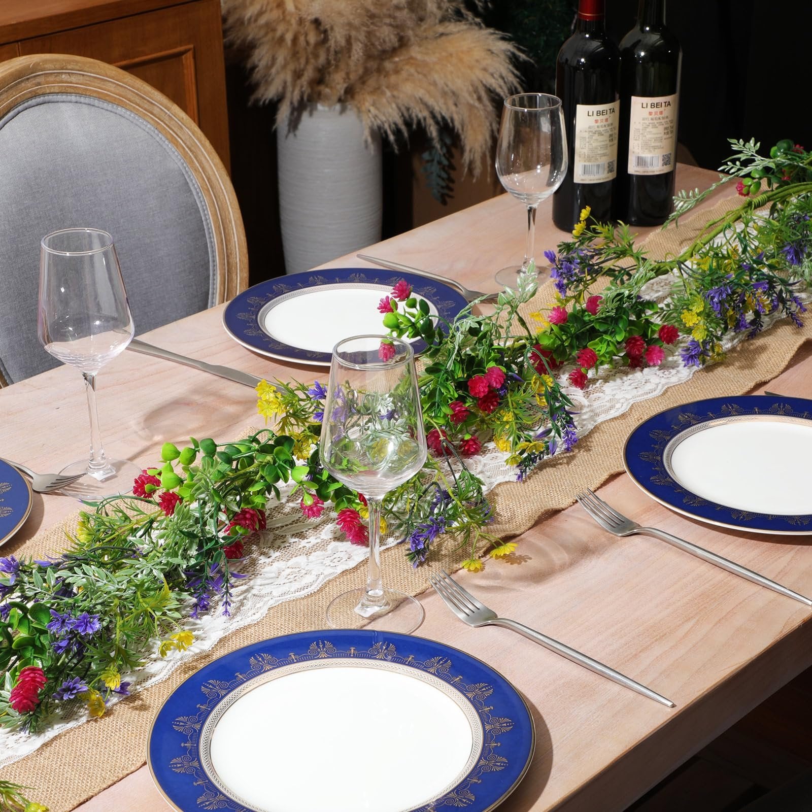 Artificial wildflower garland with daisies and lavender draped across a table setting with plates, wine glasses, and wine bottles.