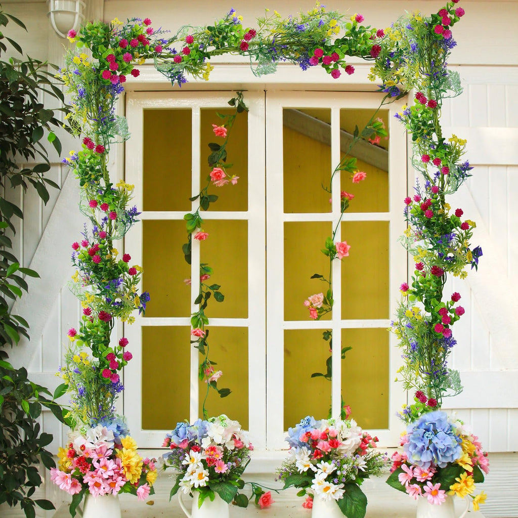 Artificial wildflower garland with pink, yellow, and purple flowers draped around a white window frame, with floral arrangements below.