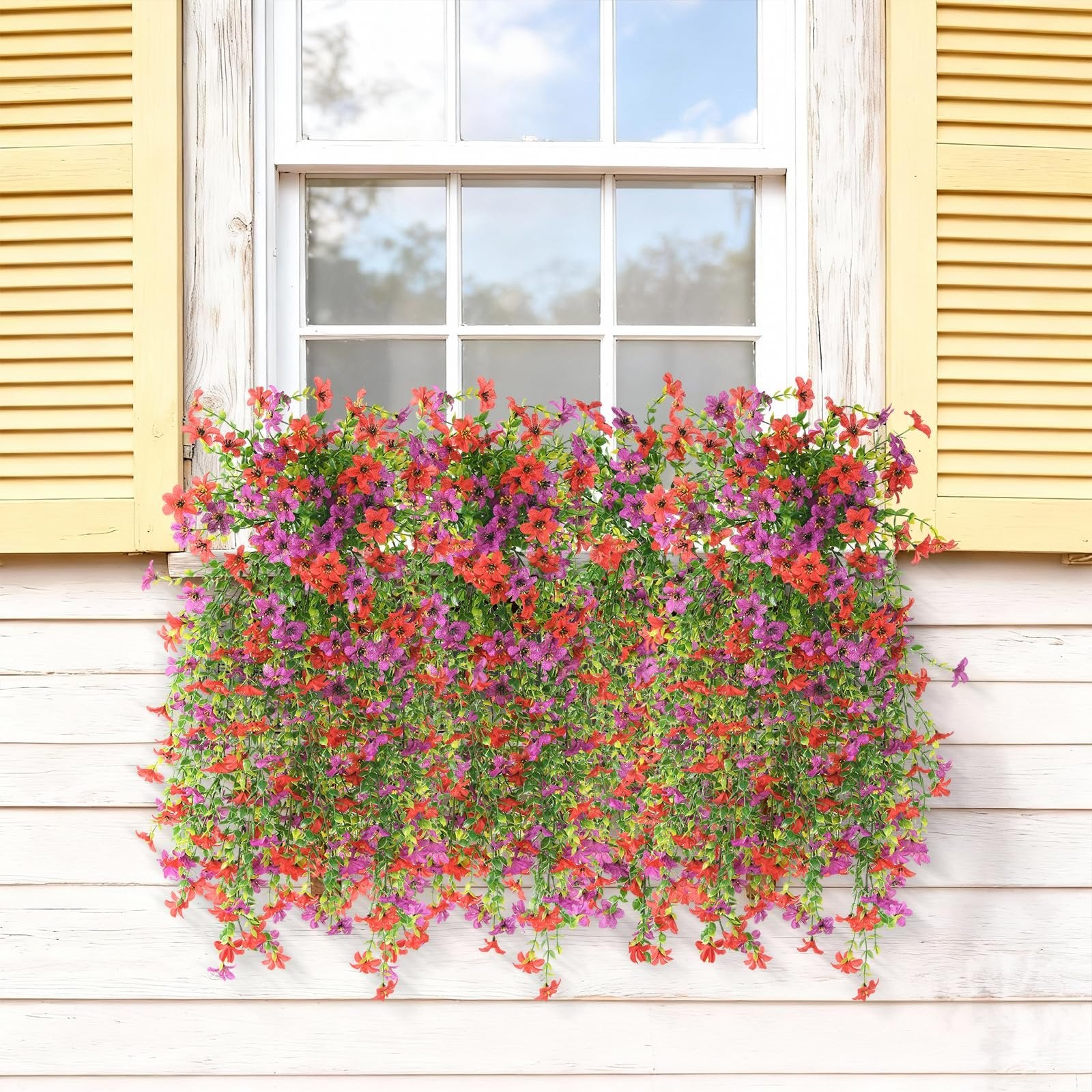 Two bunches of artificial red and purple flowers cascading down a white wooden wall next to a window with yellow shutters.
