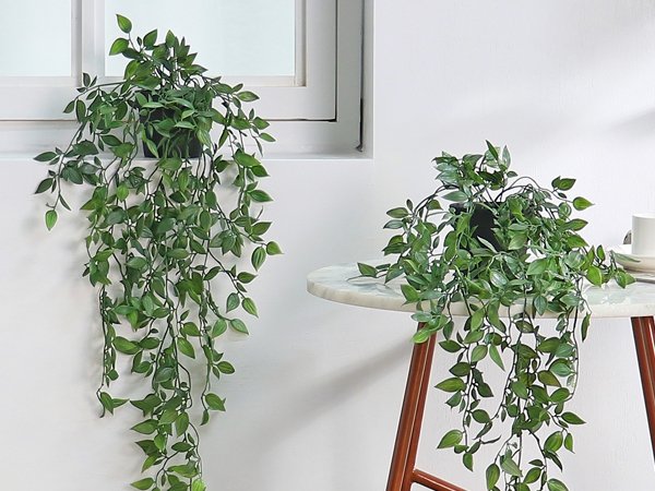 Two artificial green hanging plants in black pots, one on a windowsill and one on a marble-top table.