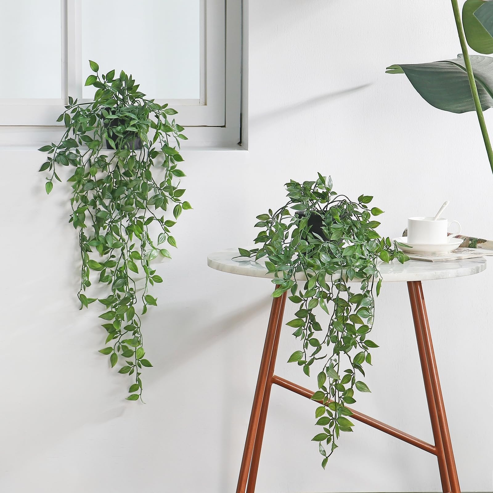 Two artificial green hanging plants, one on a windowsill and one on a side table with a cup.