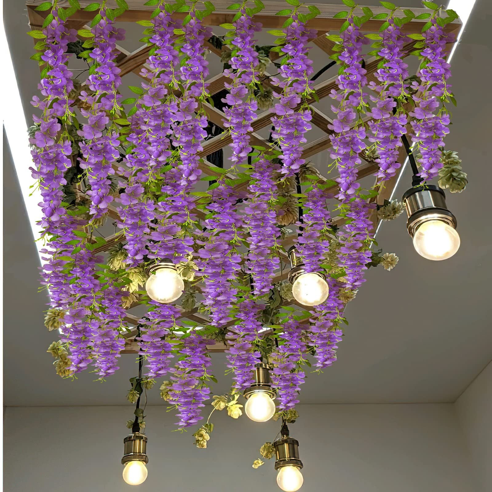Artificial purple wisteria vines with green leaves hanging from a wooden lattice, with pendant lights.