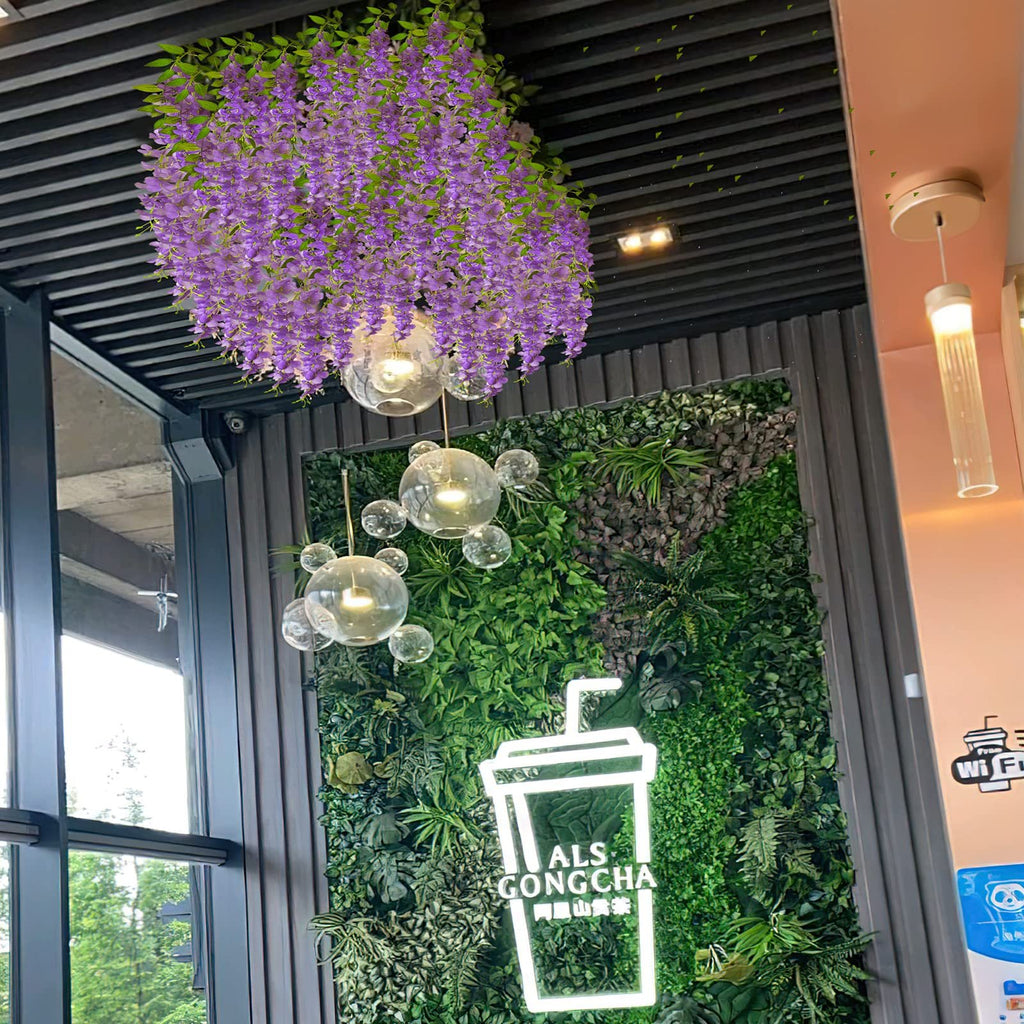 Purple artificial wisteria flowers hanging from a ceiling with decorative glass orbs and a green living wall with a neon sign.
