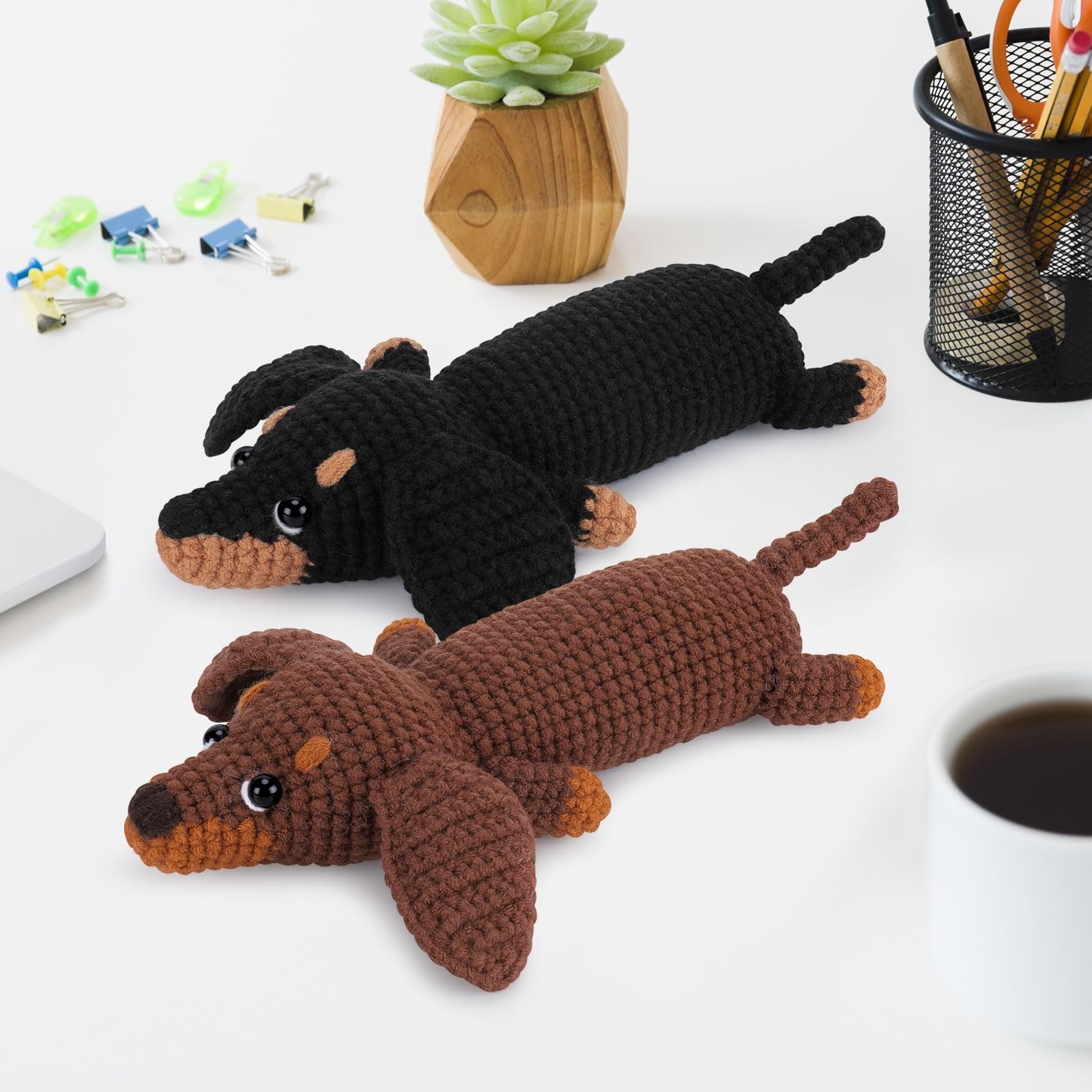 Two crocheted dachshunds, one black and one brown, lie on a white desk next to office supplies and a potted plant.