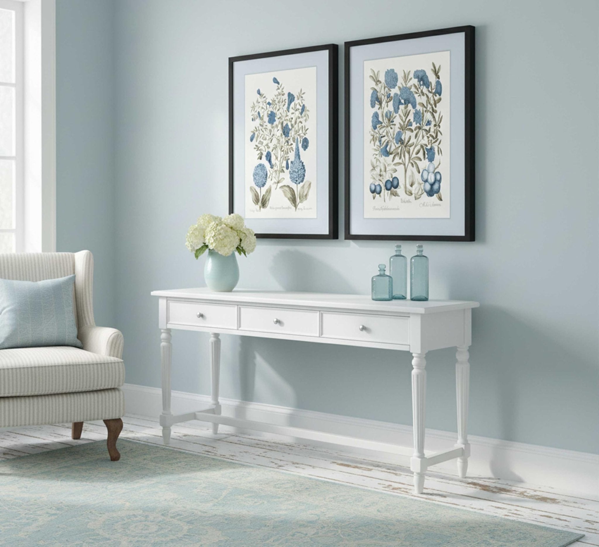 Two framed botanical prints of blue flowers hang above a white console table with decorative bottles and a vase of hydrangeas.