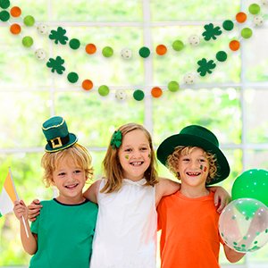 Three smiling children in St. Patrick's Day hats and face paint pose in front of two felt ball garlands with shamrocks and pom poms hanging.