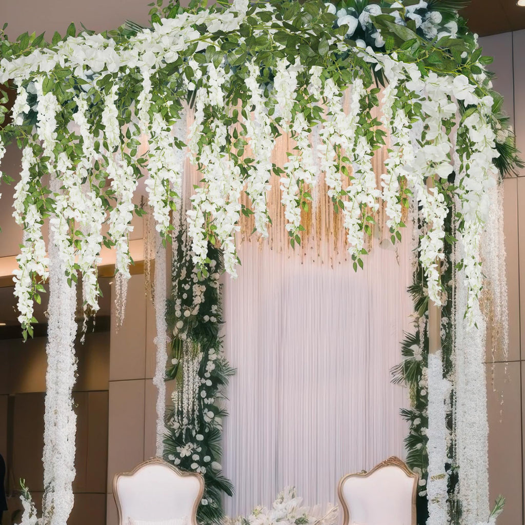 White artificial wisteria garland draped over a wedding arch with green foliage and two ornate chairs below.