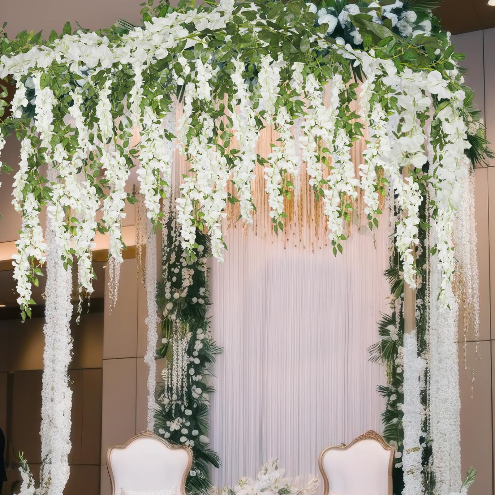 White artificial wisteria garland draped over a wedding arch with green foliage and two ornate chairs below.