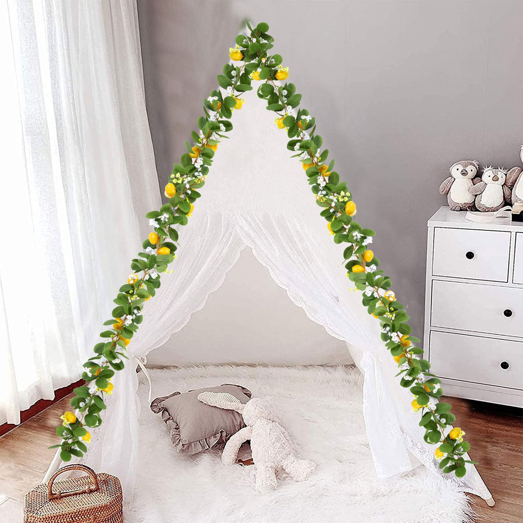 Artificial lemon vine garland with green leaves and white flowers draped over a white tent indoors.