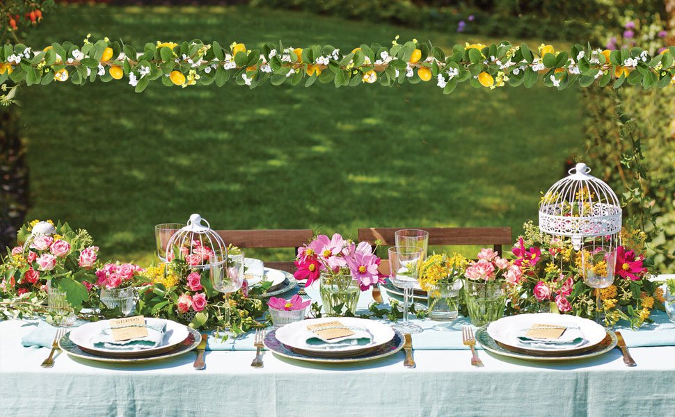 Outdoor table setting with artificial lemon garland draped above, featuring place settings and floral arrangements.