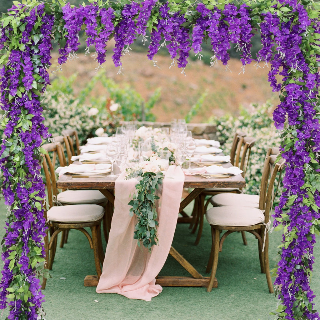 Purple wisteria garlands framing an outdoor wedding reception table setting with floral centerpieces and place settings.