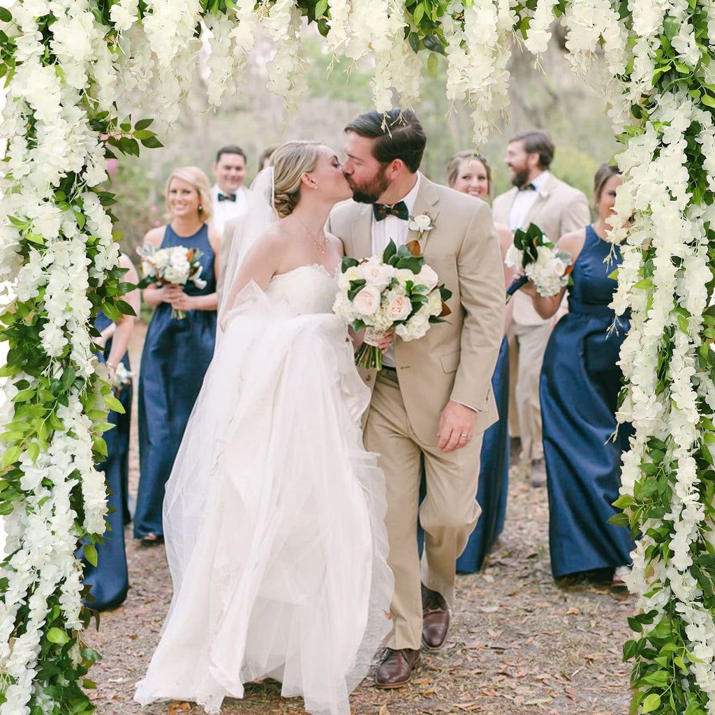 Wedding couple kissing under white wisteria flower arch with bridal party in background