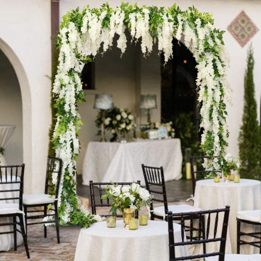 White faux wisteria garland draped over an outdoor wedding arch with tables and chairs in the background.