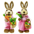 Two sisal bunny figurines in pink outfits, one holding carrots and a basket of carrots, the other holding flowers and a basket of Easter eggs.