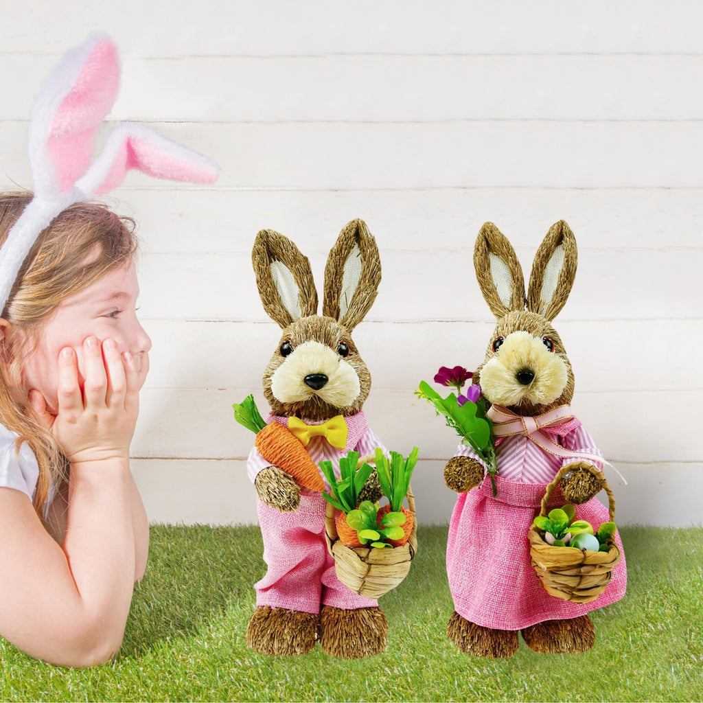 Girl wearing bunny ears looks at two straw bunny figurines holding carrots and Easter eggs on grass
