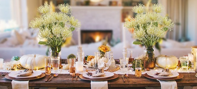 Two bouquets of pale yellow artificial chrysanthemum flowers arranged on a dining table set for a meal, with pumpkins and candles.