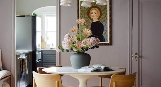 Pink artificial chrysanthemum bouquet in a vase on a table with an open book, in a dining room setting.
