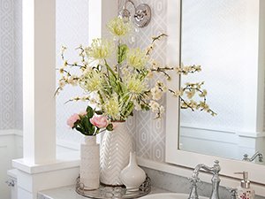 Two vases of artificial flowers, including pink chrysanthemums and white blossoms, arranged on a bathroom vanity with a mirror.