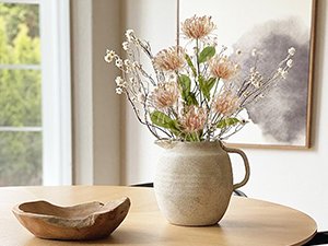 Pink artificial chrysanthemum bouquet in a ceramic pitcher on a wooden table with a wooden bowl.