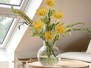 Bouquet of yellow spider mum flowers in a clear glass vase on a wooden table, with a skylight and sofa in the background.