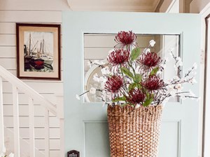 Artificial chrysanthemum bouquet in a woven basket by a light blue door with a framed picture on the wall.