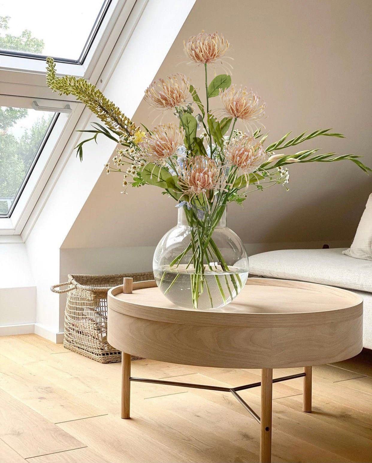Artificial pink chrysanthemum bouquet in a glass vase on a wooden table, with a skylight and basket in the background.