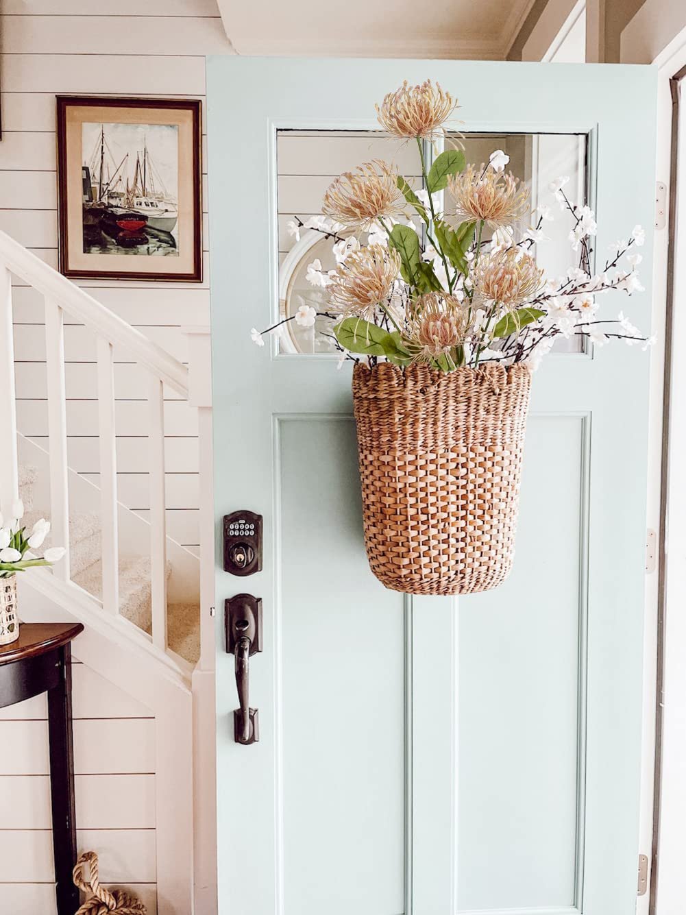 Artificial chrysanthemum bouquet in a woven basket hanging on a light blue door, with a staircase and artwork visible.
