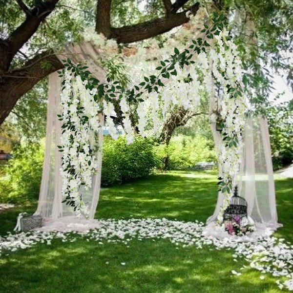 White artificial wisteria flower vines draped over a wedding arch in a garden setting with sheer fabric and scattered petals.