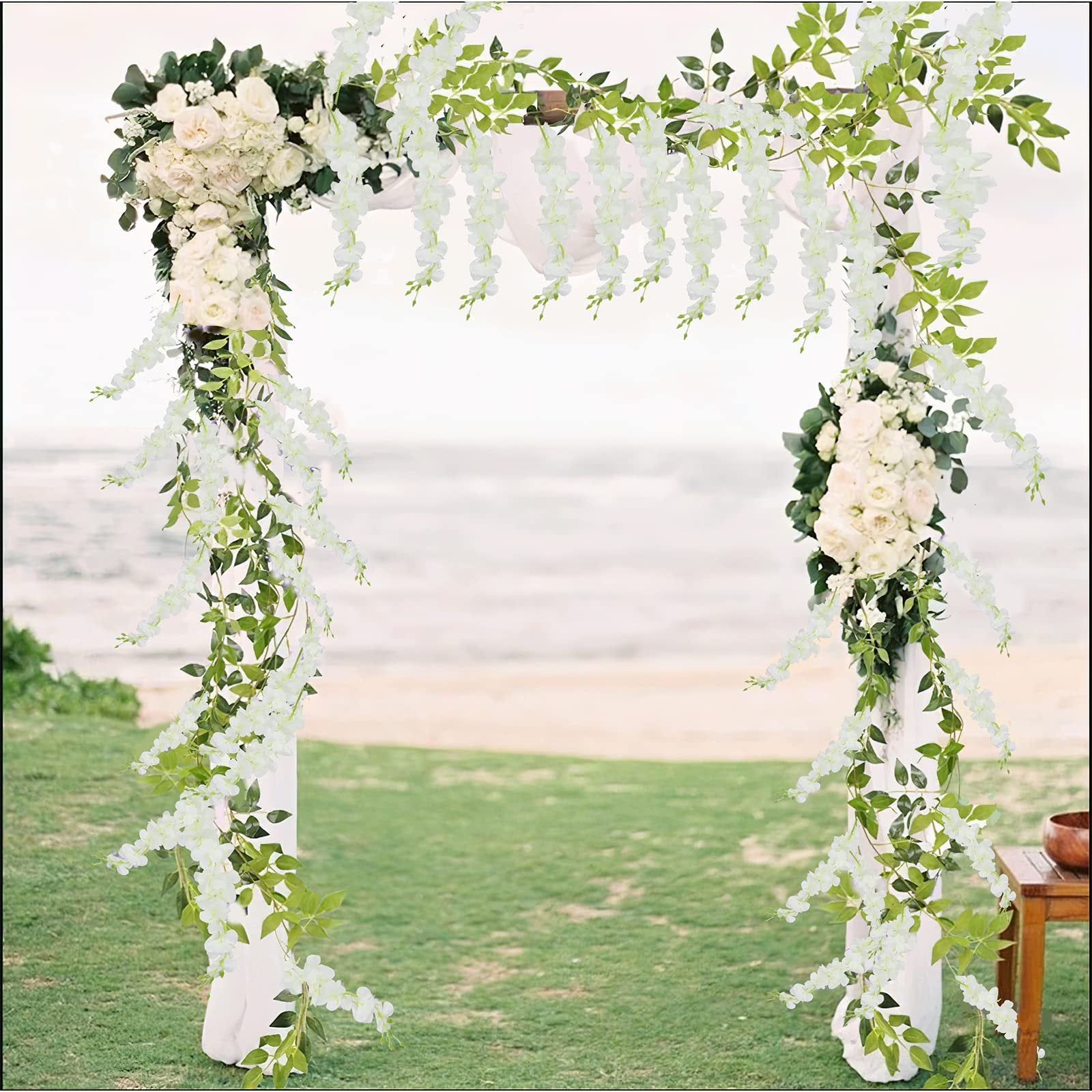 White artificial wisteria vines with green leaves draped over a wedding arch on a beach with the ocean in the background.