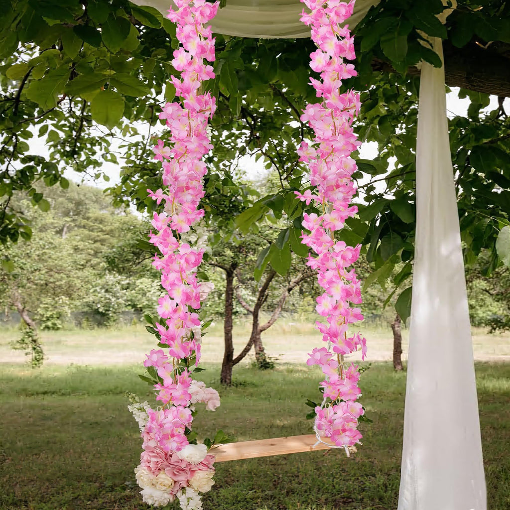 Two strands of artificial pink cherry blossom garland draped over a wooden swing seat in a garden setting with white fabric draped on the right.