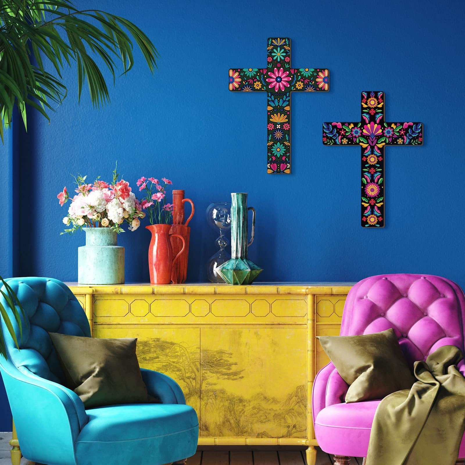 Two colorful Mexican floral wooden crosses displayed on a blue wall above a yellow cabinet with two chairs.