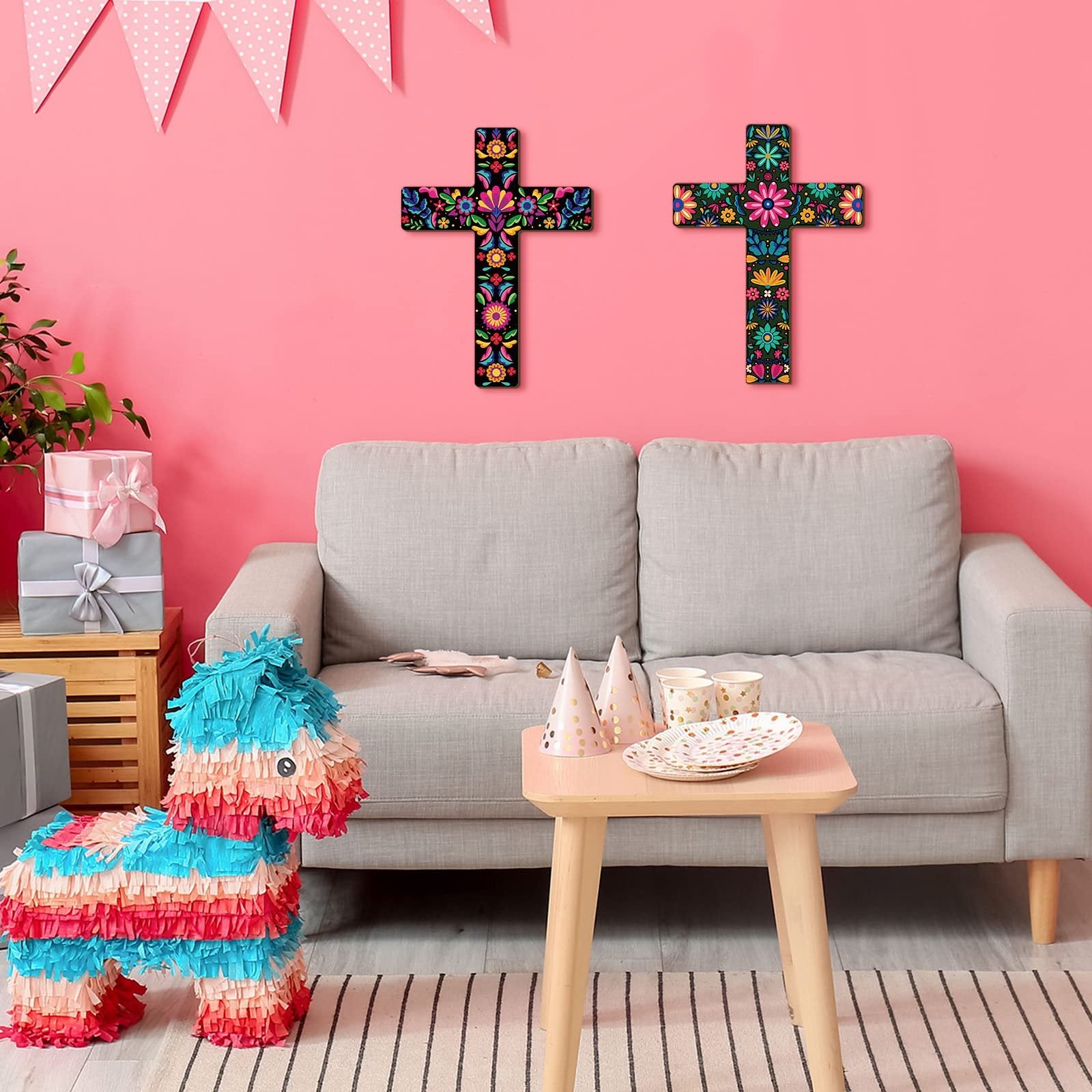 Two hand-painted floral wooden crosses hanging on a pink wall above a sofa, with a piñata and party decorations.