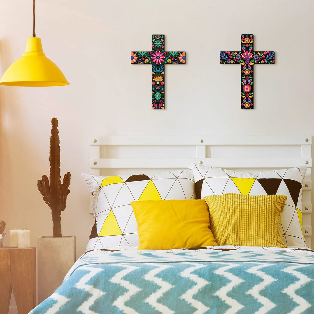 Two colorful floral wooden crosses hanging on a white wall above a bed with yellow and white pillows.
