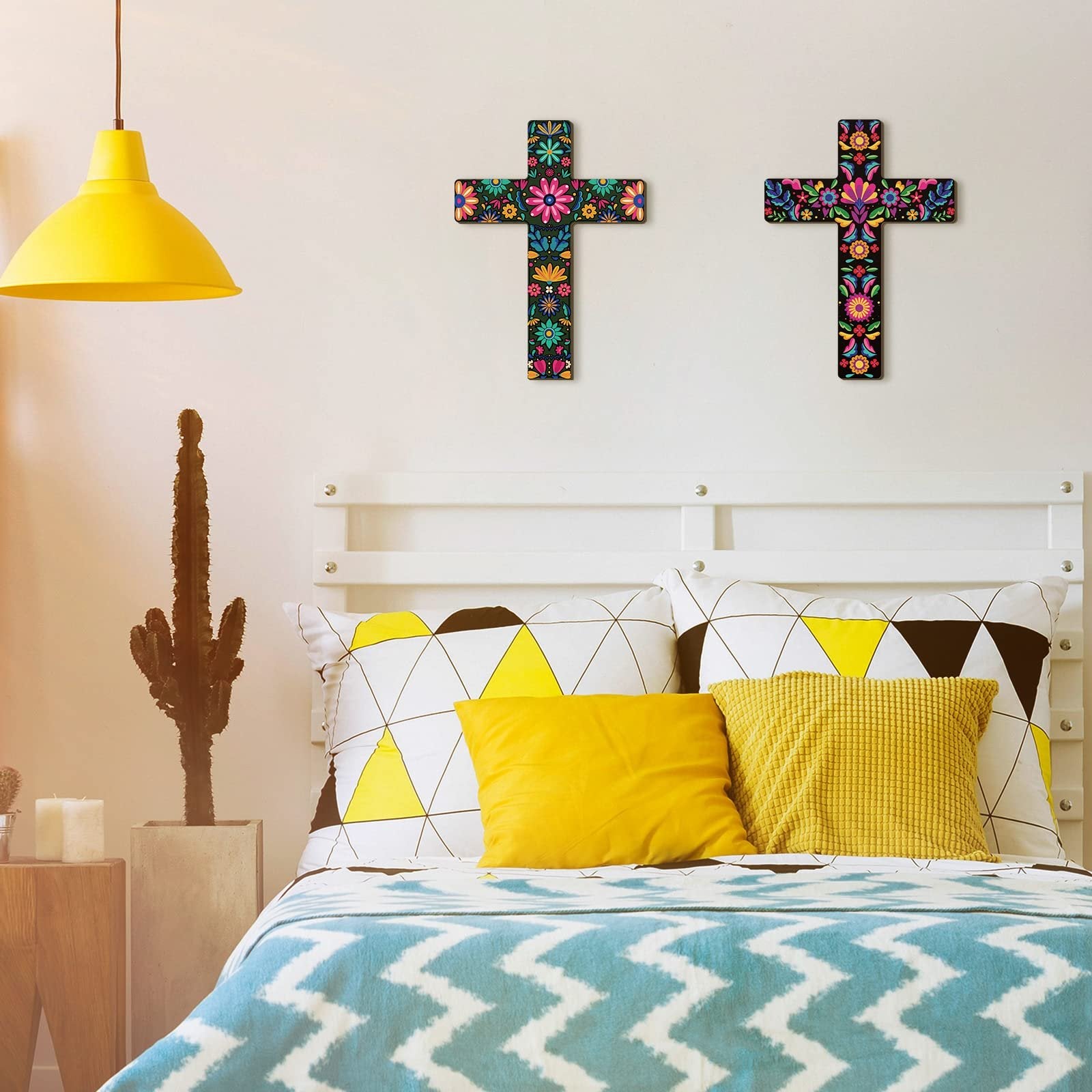 Two colorful floral wooden crosses hanging on a white wall above a bed with yellow and white pillows.