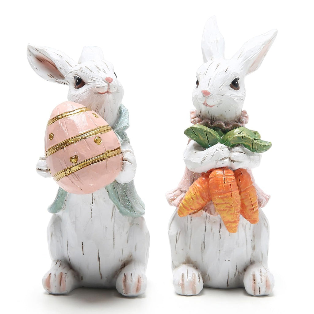 Two white resin Easter bunny figurines, one holding a pink egg, the other holding carrots, on a white background.