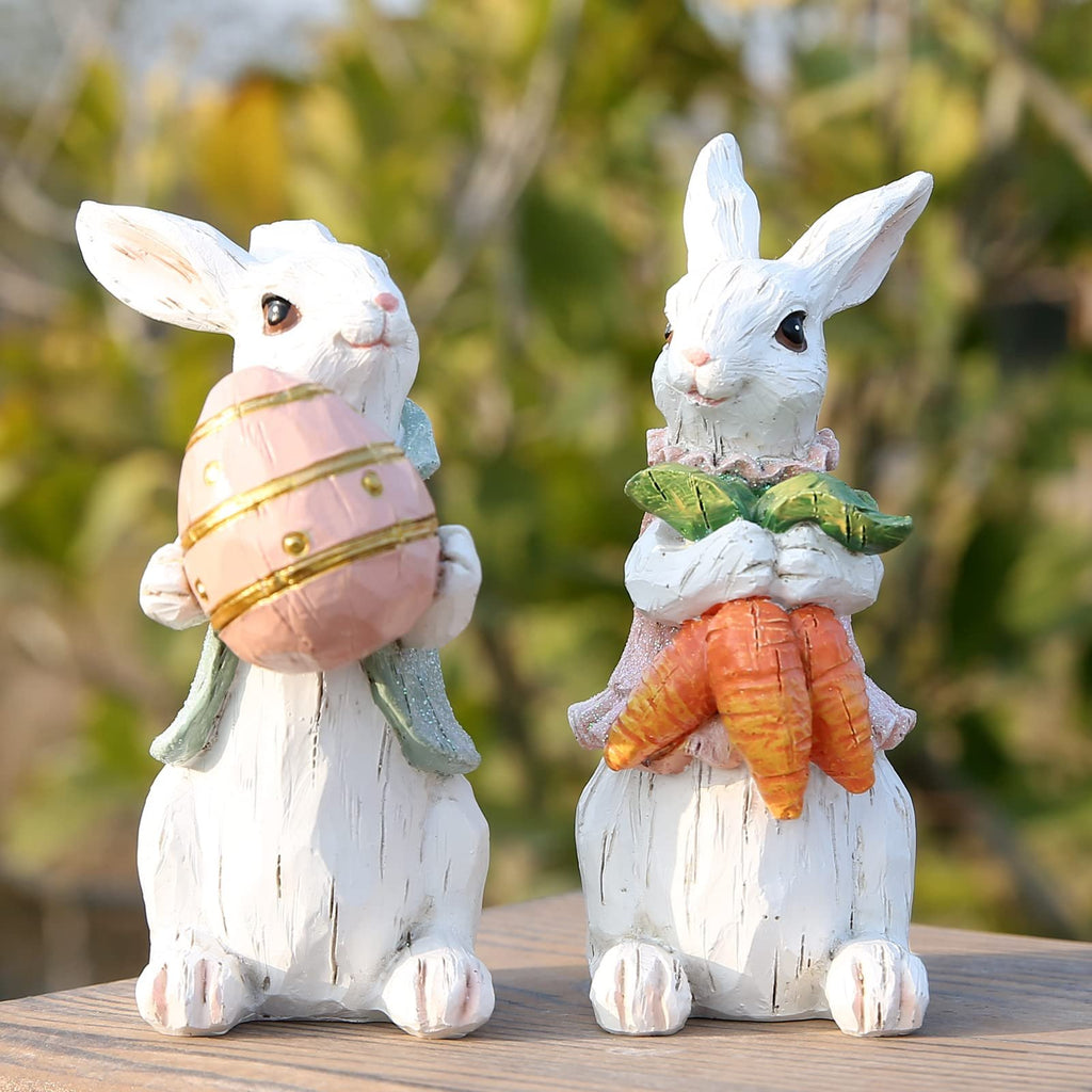 Two white resin Easter bunny figurines, one holding a pink egg, the other holding carrots, on a wooden surface.
