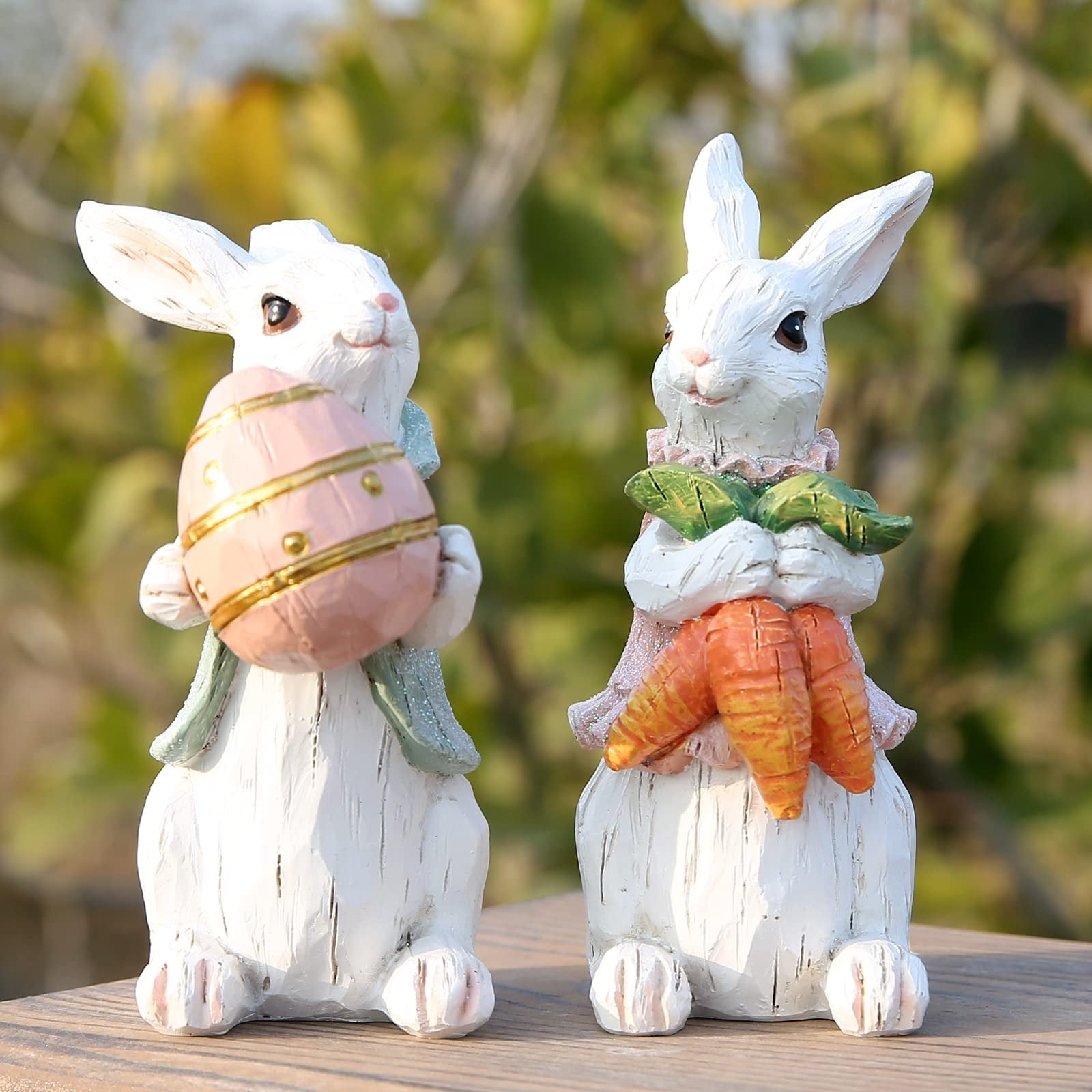 Two white resin Easter bunny figurines, one holding a pink egg, the other holding carrots, on a wooden surface.