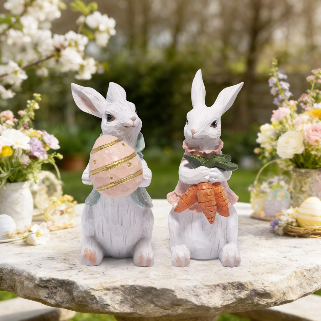 Two white resin Easter bunny figurines on a stone table, one holding a pink egg, the other holding carrots, with spring flowers in the background.