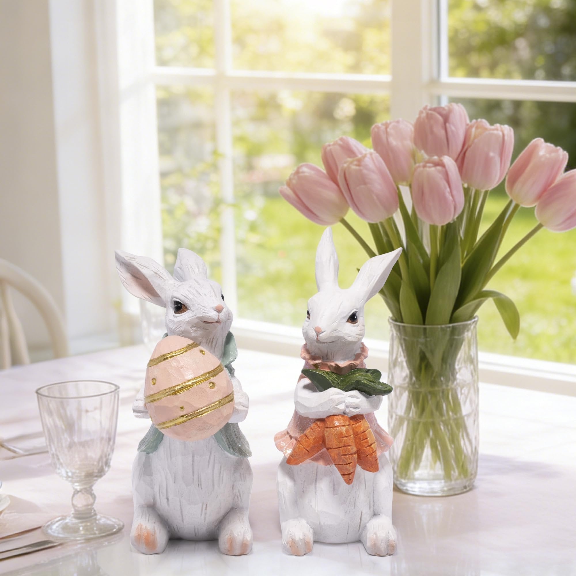 Two hand-painted resin Easter bunny figurines on a table, one holding an egg, the other carrots, with tulips in a vase.