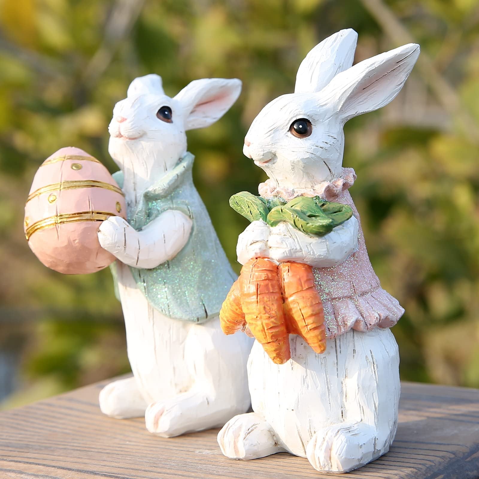 Two white resin Easter bunny figurines, one holding a pink egg, the other holding carrots, on a wooden surface with a blurred green background.